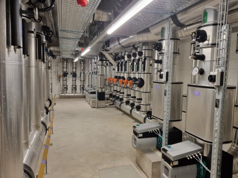 Ein Hauswirtschaftsraum im Keller mit Metallrohren, Messgeräten und Industriemaschinen. Er verfügt über komplexe Sanitär- und Elektrogeräte, darunter Bedienfelder und Ventile an den Wänden. Oben ist ein Netzwerk aus Metallleitungen sichtbar.
(Hinweis: In Ihrem bereitgestellten Text gibt es keine Vorkommen des Eszett-Zeichens, das ersetzt werden könnte.)