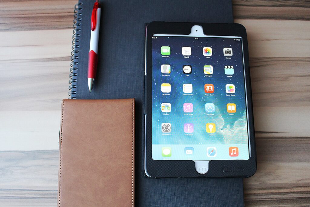 Auf einem großen Notizbuch liegt ein Tablet mit Apps, dazu ein Spiralblock und ein rot-weißer Stift. Daneben liegt eine braune Lederbrieftasche, alles auf einem Holztisch.
