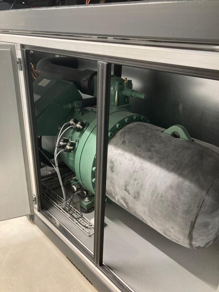 Eine große Industriemaschine in einem Metallgehäuse mit einem zylindrischen Bauteil, das mit Isolierung ummantelt ist. Das Gerät scheint Teil eines Kühl- oder HLK-Systems zu sein, Rohre und Armaturen sind sichtbar.