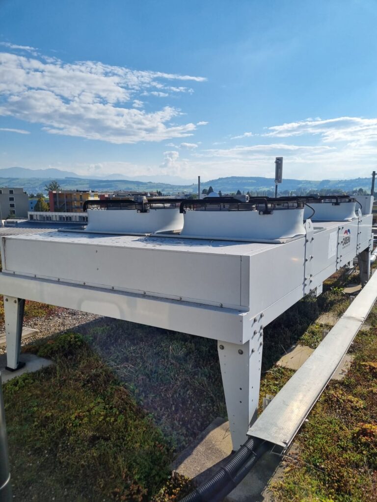 Das Bild zeigt eine große industrielle Klimaanlage auf einem Dach vor einem klaren blauen Himmel mit vereinzelten Wolken. Im Hintergrund sind Hügel und entfernte Gebäude im hellen Sonnenlicht zu sehen.