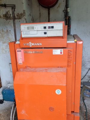 Alter orangefarbener Viessmann Vitola-Biferral-Kessel mit Bedienfeld, aufgestellt in einem abgenutzten Raum mit Rohren und einer roten Sicherheitsvorrichtung an der Wand. Rost und Schmutz am Gerät und in der Umgebung sichtbar.