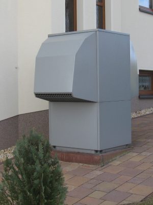 Eine große, graue Wärmepumpeneinheit für den Außenbereich, die neben einem Gebäude auf einem gepflasterten Gehweg installiert ist. Im Vordergrund ist ein kleiner Strauch zu sehen.