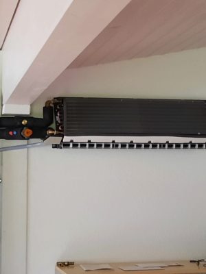 Eine an der Wand montierte Klimaanlage mit freiliegenden Rohren in einem Raum. Darunter steht ein Tisch mit ein paar Flaschen, darunter eine Flasche Rotwein, und einigen Papieren. Die Decke ist aus Holz mit sichtbaren Balken.

(Hinweis: Das Eszett-Zeichen kommt im angegebenen Text nicht vor.)