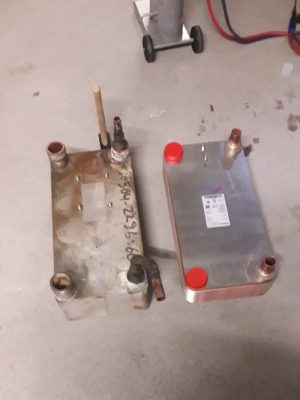 Zwei Wärmetauscher, ein alter und ein neuer, stehen nebeneinander auf einem Betonboden. Der alte Wärmetauscher sieht rostig und abgenutzt aus, während der neue sauber und glänzend ist und rote Kappen an den Anschlüssen hat.