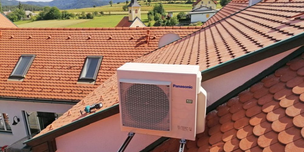 Der Text enthält kein Eszett-Zeichen und bleibt daher unverändert: Eine Klimaanlage von Panasonic ist seitlich an einem roten Ziegeldach montiert. Im Hintergrund sind ein Kirchturm und eine malerische Landschaft mit Bäumen und Feldern unter einem teilweise bewölkten Himmel zu sehen.