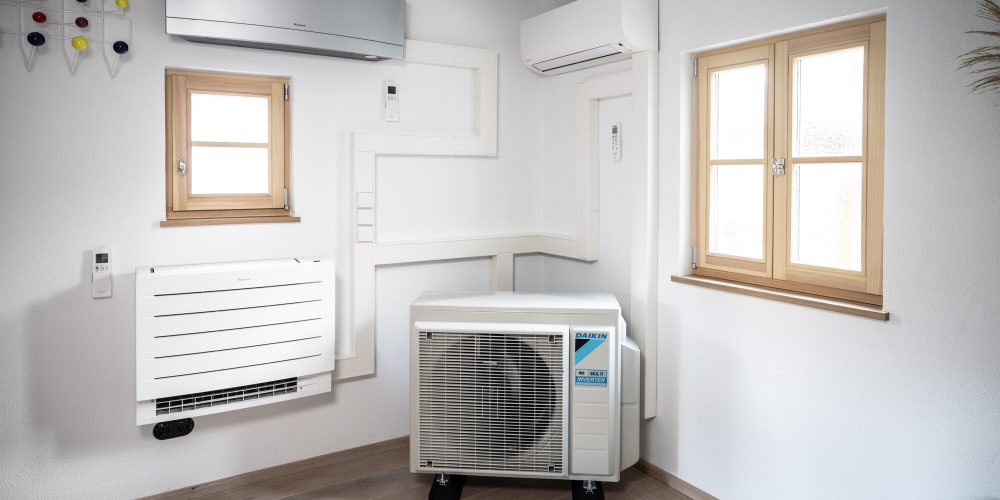 Ein moderner Raum mit zwei kleinen Fenstern, einer an der Wand montierten Klimaanlage, einer Luftaufbereitungsanlage für den Innenbereich und einer HLK-Anlage für den Außenbereich neben einer weißen Wand mit Akzenten aus Holzbalken. Ein dekoratives Wandstück mit bunten Knöpfen ist ebenfalls sichtbar.